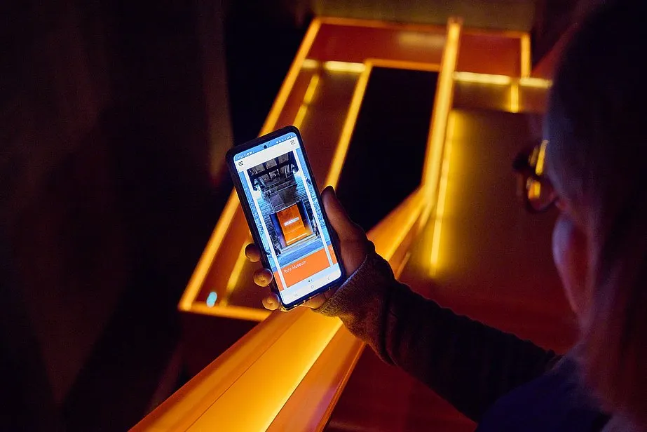 Eine Person hält ein Smartphone in der rechten Hand und fotografiert oder betrachtet ein beleuchtetes Treppenhaus mit orangefarbenen Lichtstreifen. Die Szene ist in dunkler Umgebung aufgenommen, die Lichtakzente heben die geometrischen Formen der Treppe hervor. Auf dem Smartphone-Display ist das gleiche Motiv wie im Hintergrund zu sehen.