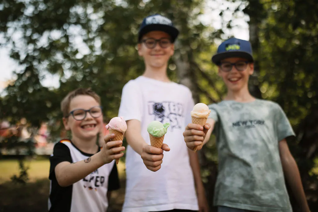 Drei lächelnde Kinder stehen im Freien unter Bäumen und halten ihre Eiswaffeln in Richtung Kamera. Im Vordergrund sind die Eiskugeln in Rosa, Grün und Hellgelb scharf zu sehen, während die Gesichter der Kinder leicht unscharf bleiben. Die Szene wirkt sommerlich, fröhlich und unbeschwert.