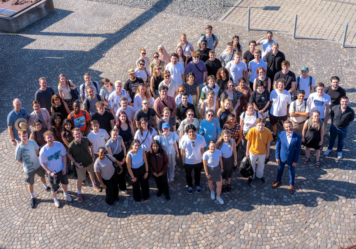 Große Gruppe von Menschen steht auf einem gepflasterten Platz und blickt nach oben in die Kamera. Die Aufnahme ist aus erhöhter Perspektive gemacht. Die Personen stehen dicht beieinander in mehreren Reihen, viele lächeln. Auf dem Platz sind lange Schatten zu sehen, die auf helles Sonnenlicht hinweisen.