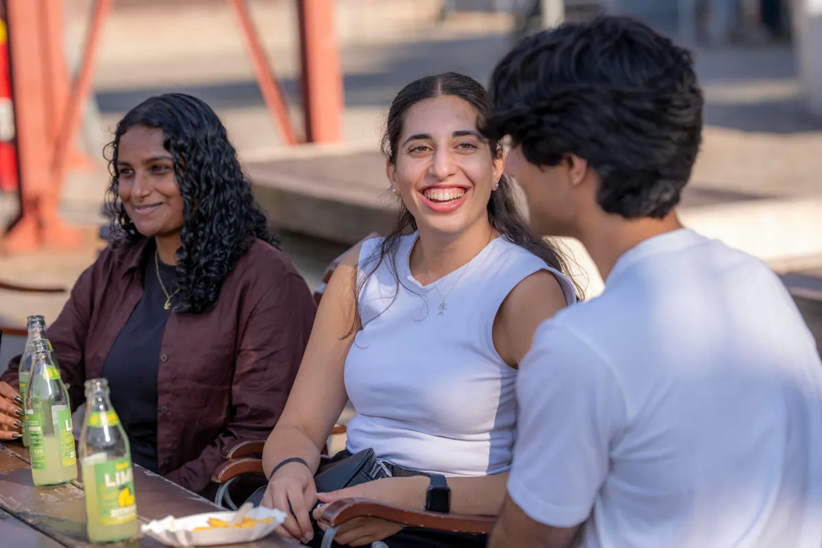 Drei junge Erwachsene sitzen draußen an einem Tisch und unterhalten sich. In der Mitte lacht eine Frau in einem hellen ärmellosen Oberteil und blickt zu einem Mann rechts im Bild. Links sitzt eine weitere Frau mit lockigem Haar. Auf dem Tisch stehen Getränkeflaschen und eine kleine Schale. Im Hintergrund sind unscharf rote Stahlstützen und Gebäudeteile zu sehen.