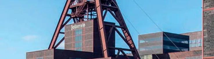 Ausschnitt des Förderturms und der Backsteingebäude der Zeche Zollverein vor blauem Himmel. Im Vordergrund stehen dunkle, kubische Industriegebäude mit großen Fensterflächen, darüber erhebt sich die markante rotbraune Stahlkonstruktion des Fördergerüsts.