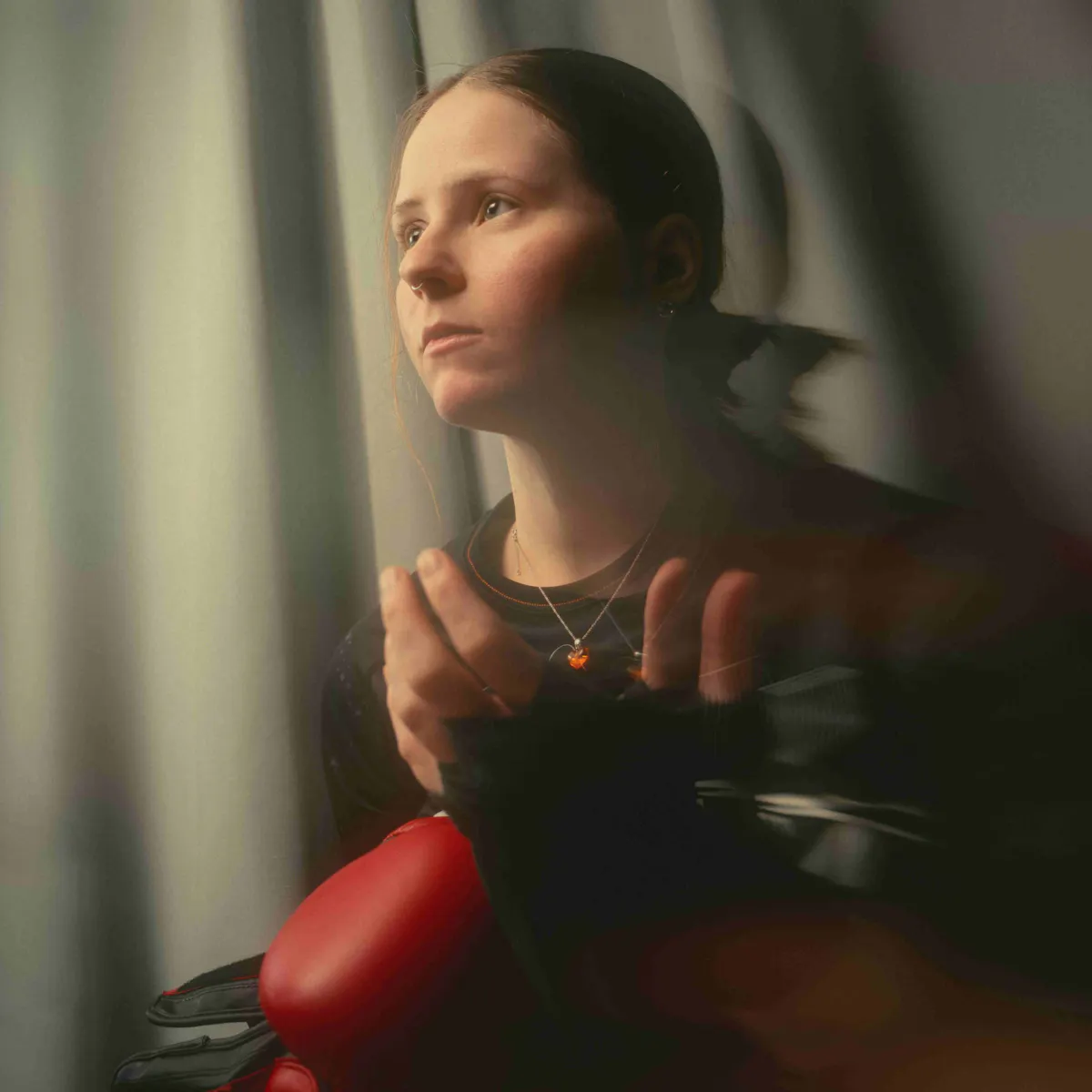 Eine junge Frau mit zusammengebundenen Haaren steht vor einem hellen Vorhang und blickt nachdenklich zur Seite. Sie trägt ein dunkles Oberteil, eine Halskette mit einem kleinen Anhänger und hält einen roten Boxhandschuh in der Hand. Die Szene ist weich ausgeleuchtet, mit verschwommenen Bewegungsunschärfen im Vordergrund, was eine verträumte, poetische Stimmung erzeugt.