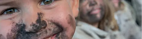 Nahaufnahme eines lächelnden Kindes mit verschmutztem Gesicht, das direkt in die Kamera blickt. Im unscharfen Hintergrund sitzen weitere Kinder oder Jugendliche, deren Gesichter ebenfalls mit dunklen Flecken verschmiert sind.