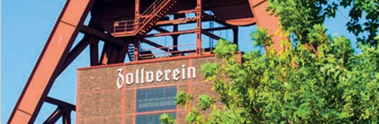 Ausschnitt des Förderturms der Zeche Zollverein vor blauem Himmel. Unter der rotbraunen Stahlkonstruktion ist ein Backsteingebäude mit dem Schriftzug Zollverein zu sehen, teilweise verdeckt von grünen Baumkronen im Vordergrund.