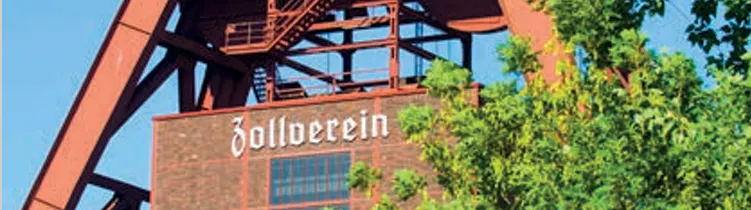 Ausschnitt des Förderturms der Zeche Zollverein vor blauem Himmel. Unter der rotbraunen Stahlkonstruktion ist ein Backsteingebäude mit dem Schriftzug Zollverein zu sehen, teilweise verdeckt von grünen Baumkronen im Vordergrund.