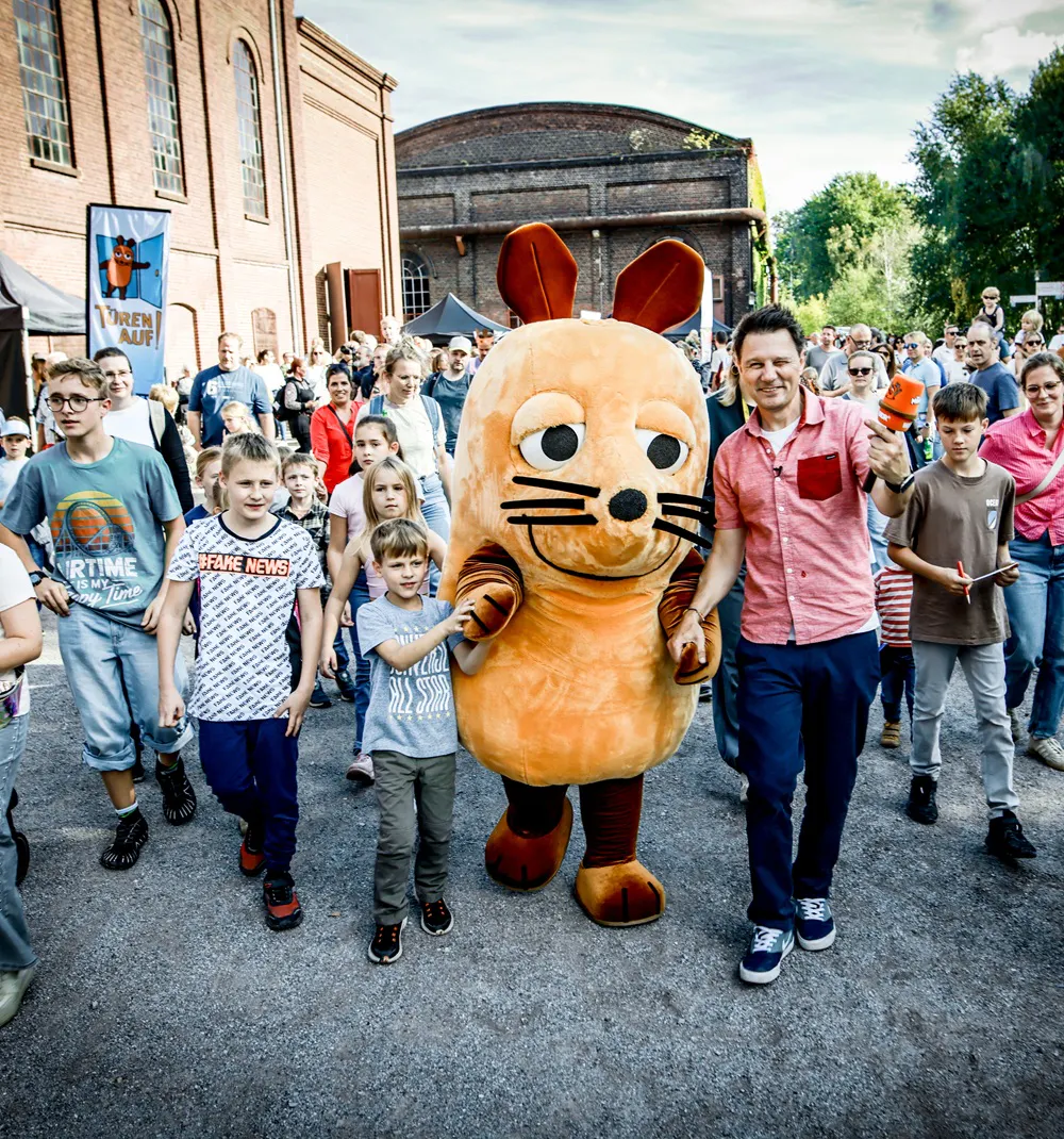 Menschenmenge bei einer Veranstaltung im Freien vor historischen Backsteingebäuden. In der Mitte geht eine Person im Mauskostüm zwischen Kindern und Erwachsenen. Rechts daneben hält ein Mann ein orangefarbenes Mikrofon. Mehrere Kinder laufen im Vordergrund mit, im Hintergrund stehen weitere Besucherinnen und Besucher.